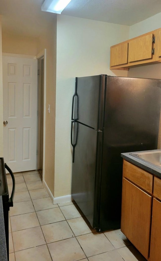 Kitchen with freestanding refrigerator, light tile patterned flooring, dark countertops, baseboards, and a sink