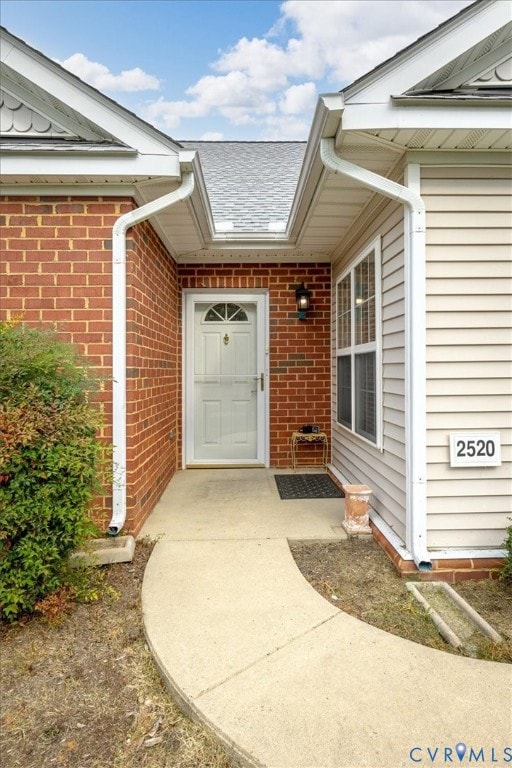 View of exterior entry with a shingled roof and brick siding