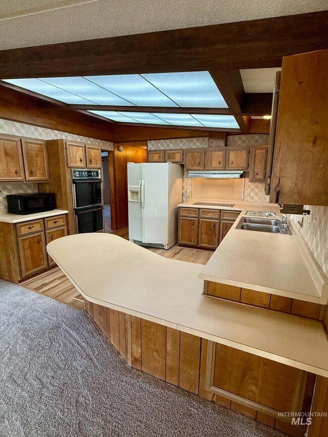 Kitchen featuring black appliances, brown cabinetry, beamed ceiling, a peninsula, and light countertops