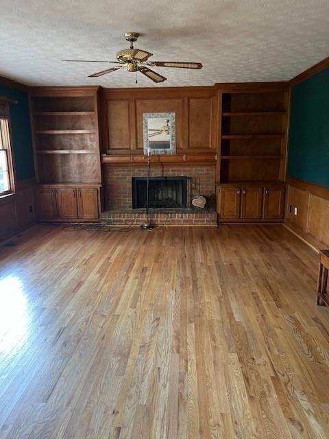 Unfurnished living room featuring built in shelves, light wood-type flooring, a brick fireplace, a textured ceiling, and a wainscoted wall