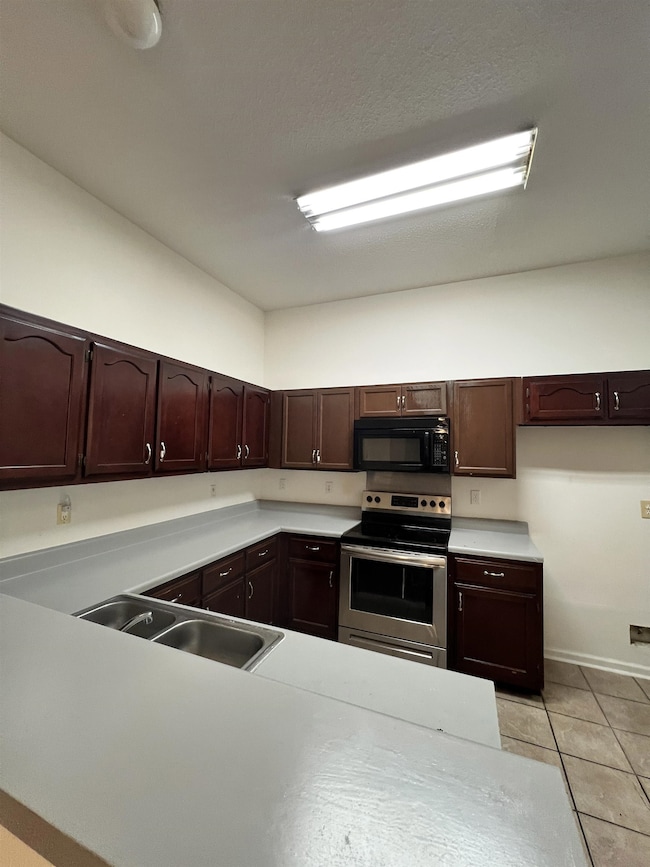 Kitchen with electric range, light tile patterned floors, light countertops, black microwave, and dark brown cabinets