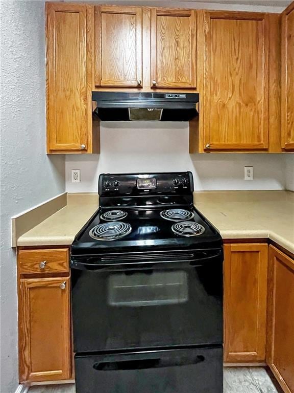 Kitchen featuring black range with electric stovetop, under cabinet range hood, light countertops, a textured wall, and brown cabinetry