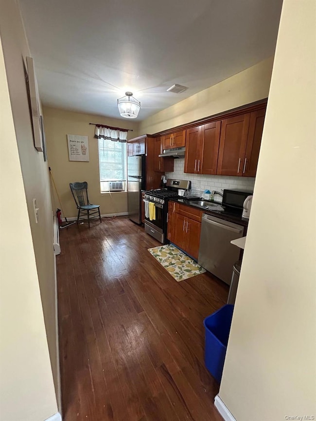 Kitchen with appliances with stainless steel finishes, dark wood finished floors, backsplash, under cabinet range hood, and brown cabinets