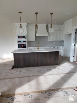 Kitchen with a center island with sink, hanging light fixtures, stainless steel appliances, white cabinets, and custom range hood