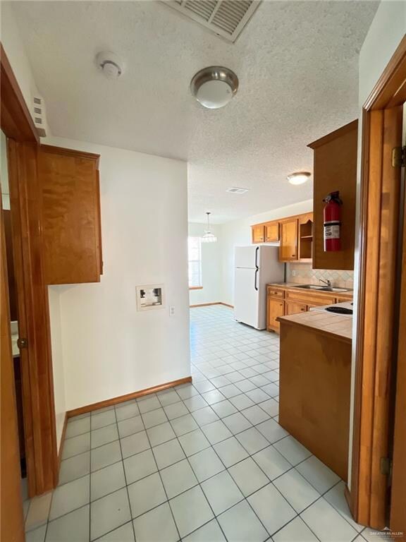 Kitchen with brown cabinetry, freestanding refrigerator, light tile patterned floors, decorative backsplash, and a textured ceiling