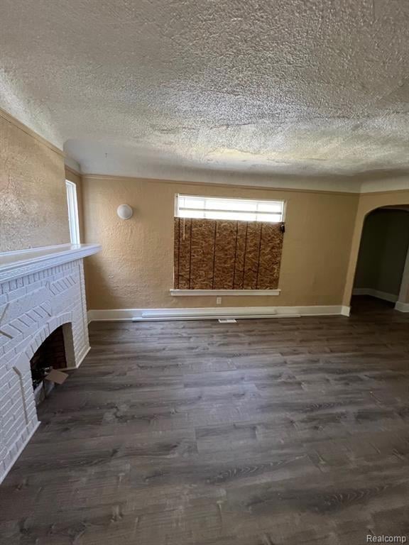 Unfurnished living room featuring a textured wall, arched walkways, dark wood-style flooring, a textured ceiling, and a brick fireplace