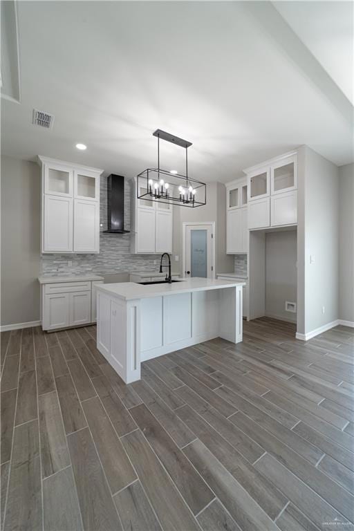 Kitchen featuring wall chimney exhaust hood, a sink, dark wood-style flooring, tasteful backsplash, and a kitchen island with sink