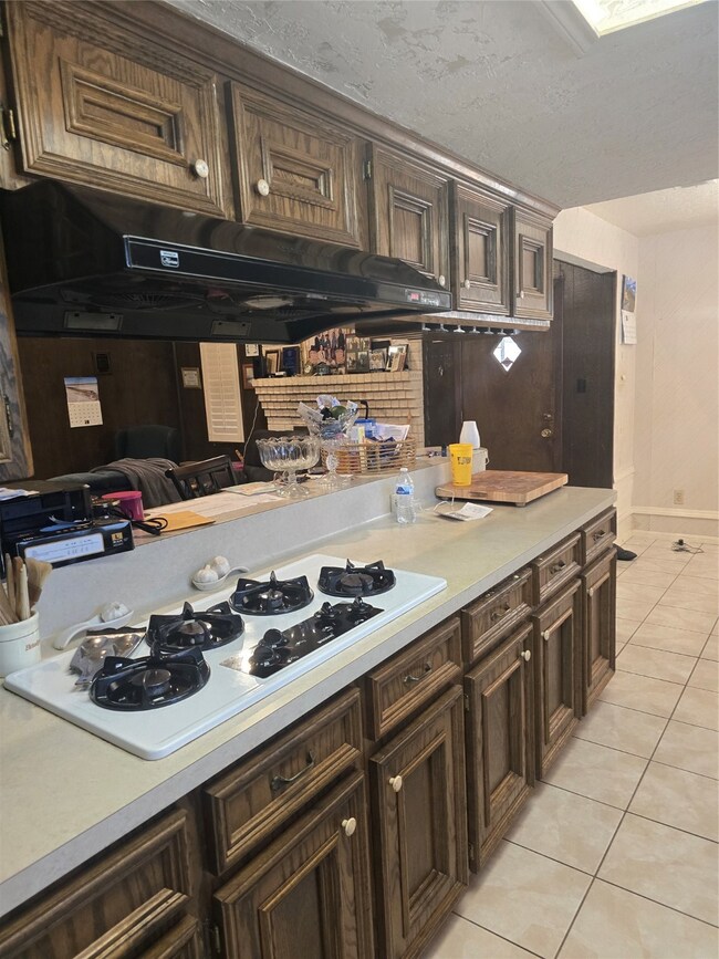 This kitchen features wooden cabinetry with a classic design, a gas cooktop, and a light-colored countertop. The space includes tiled flooring and a practical layout, ideal for cooking and meal preparation.
