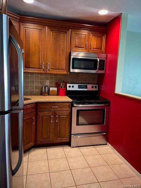 Kitchen featuring appliances with stainless steel finishes, light countertops, light tile patterned floors, backsplash, and brown cabinets