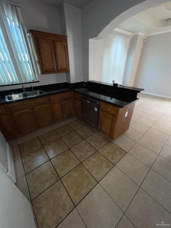Kitchen with a peninsula, brown cabinets, light tile patterned flooring, dishwashing machine, and dark stone counters