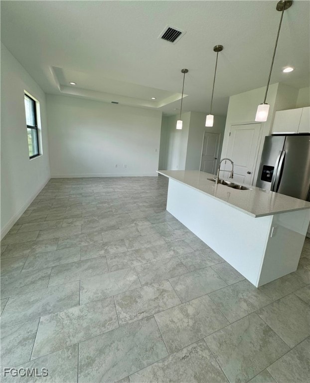 Kitchen with a raised ceiling, a center island with sink, stainless steel refrigerator with ice dispenser, light stone countertops, and white cabinets