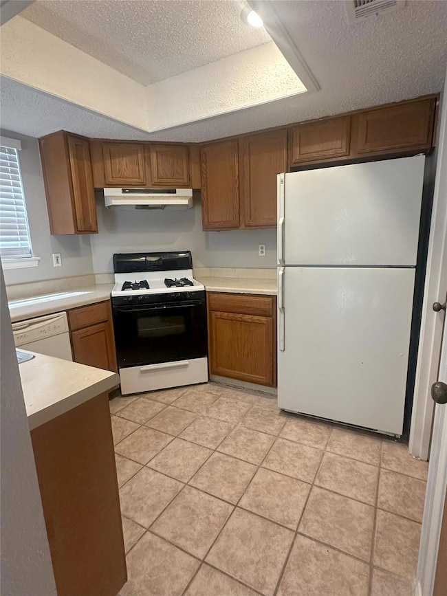 Kitchen featuring white appliances, under cabinet range hood, brown cabinetry, light countertops, and a textured ceiling