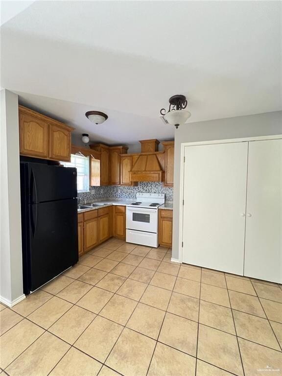 Kitchen with white electric range oven, freestanding refrigerator, custom range hood, light tile patterned flooring, and backsplash