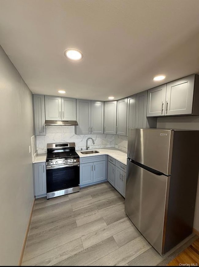 Kitchen featuring stainless steel appliances, decorative backsplash, gray cabinets, light wood-type flooring, and light stone counters
