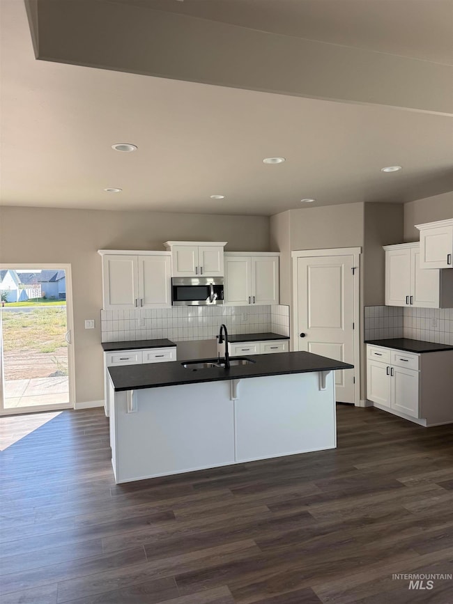 Kitchen with dark countertops, a center island with sink, stainless steel microwave, white cabinetry, and dark wood finished floors