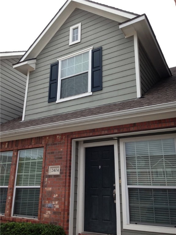View of exterior entry with brick siding and roof with shingles