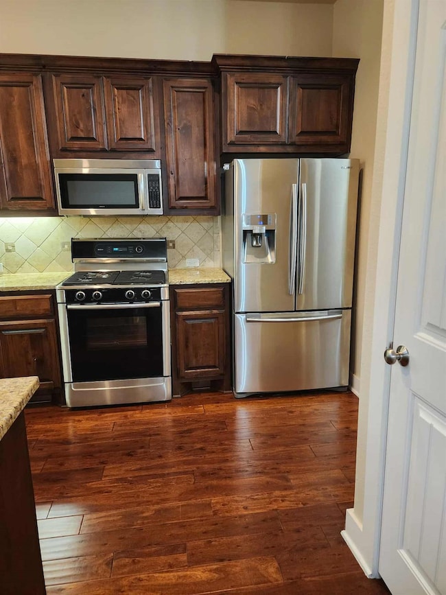 Kitchen featuring appliances with stainless steel finishes, light stone countertops, backsplash, dark brown cabinetry, and dark wood-style floors