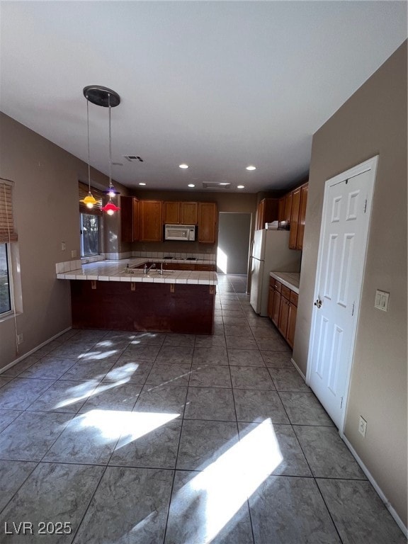 Kitchen featuring brown cabinets, a peninsula, tile counters, decorative light fixtures, and refrigerator
