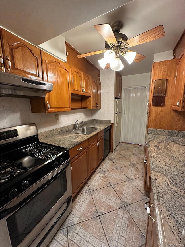 Kitchen featuring gas range, brown cabinetry, under cabinet range hood, a ceiling fan, and dark stone countertops