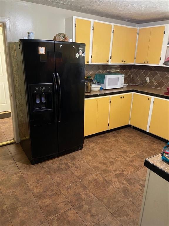 Kitchen with a textured ceiling, black fridge with ice dispenser, dark countertops, cream cabinets, and backsplash