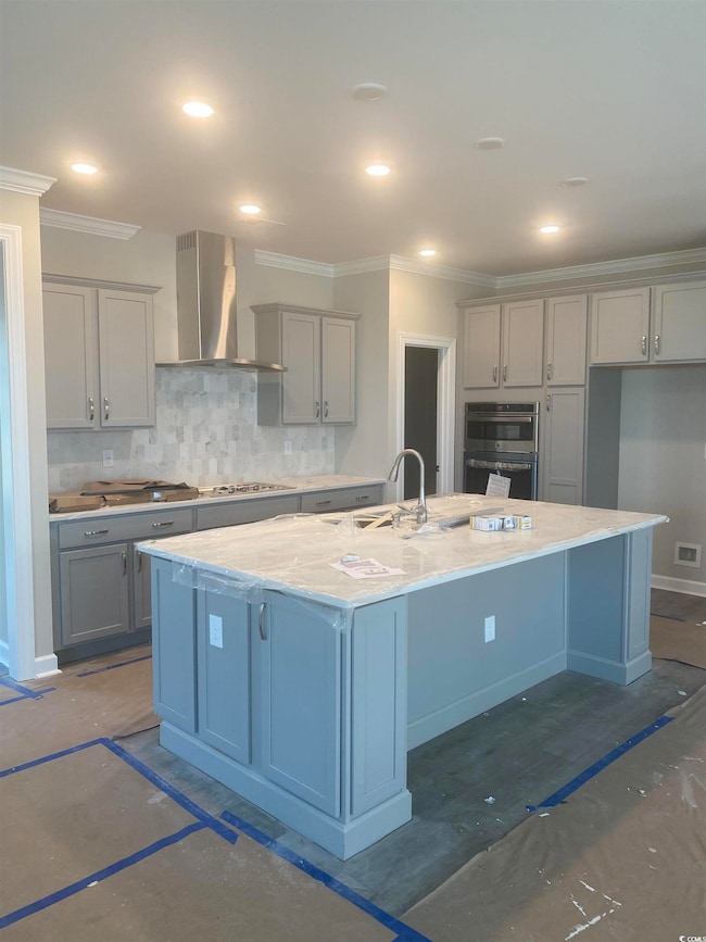Kitchen featuring wall chimney exhaust hood, gray cabinetry, an island with sink, crown molding, and decorative backsplash