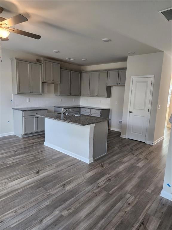 Kitchen featuring gray cabinets, ceiling fan, an island with sink, dark stone countertops, and dark wood-style flooring