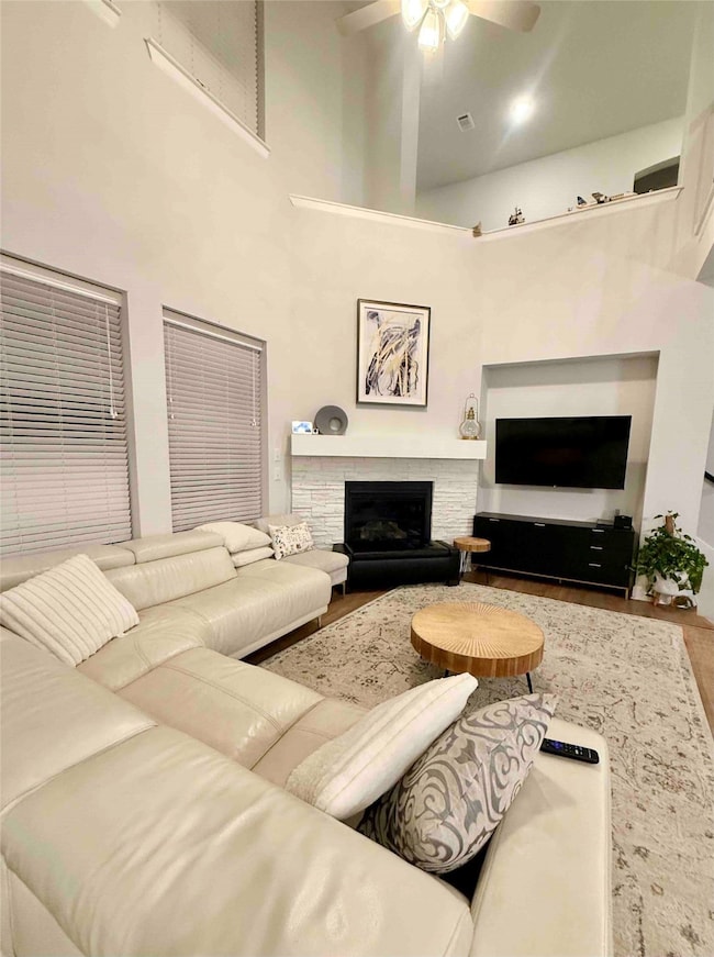 Living area with a high ceiling, a fireplace with raised hearth, wood finished floors, and a ceiling fan