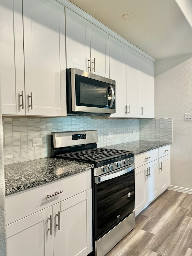 Kitchen with appliances with stainless steel finishes, white cabinetry, dark stone counters, light wood-type flooring, and backsplash