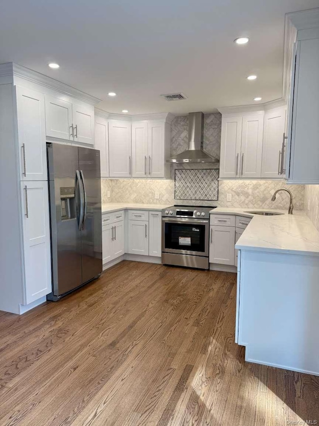 Kitchen with light stone countertops, stainless steel appliances, wall chimney exhaust hood, white cabinetry, and backsplash