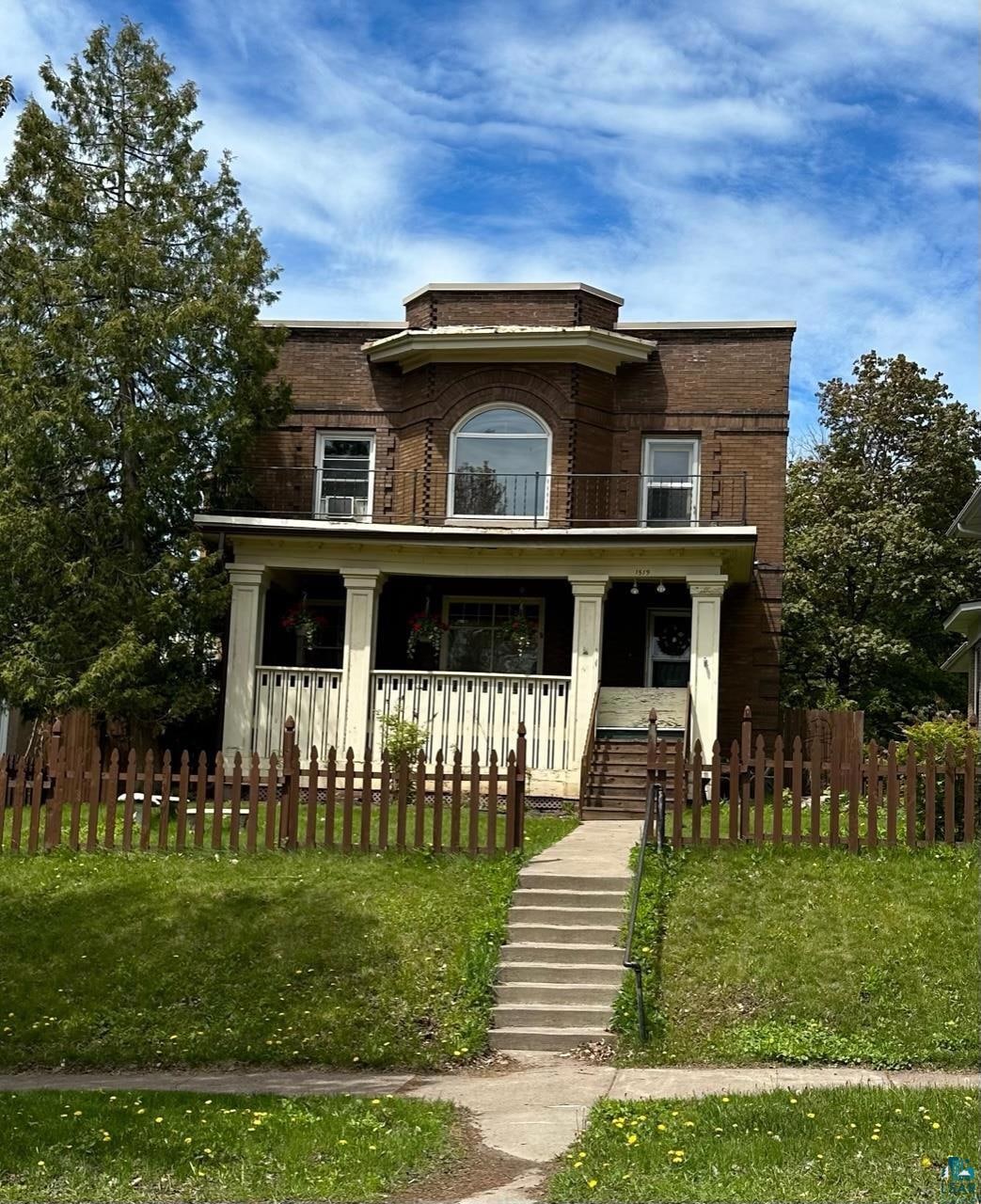 Front of property featuring a porch, a balcony, a fenced front yard, and brick siding