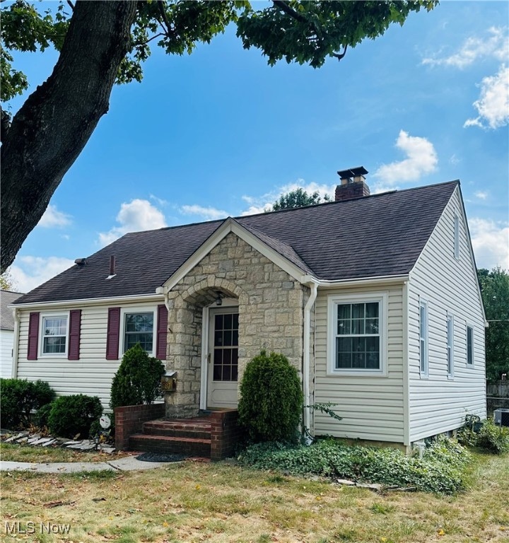 View of front of house featuring a chimney, stone siding, a front lawn, and roof with shingles
