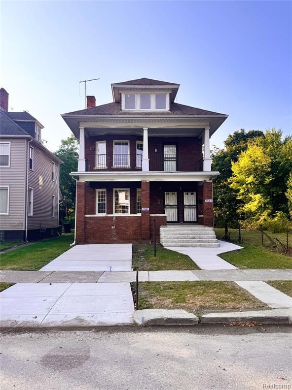American foursquare style home featuring brick siding, a balcony, a chimney, covered porch, and a front yard
