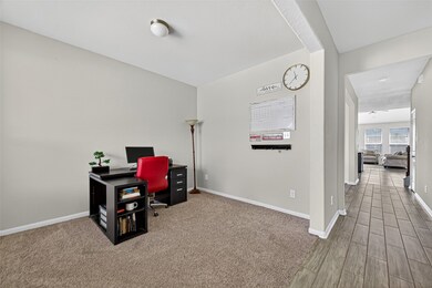 This photo showcases a home office with neutral walls and carpet flooring. It features a sleek desk with a red chair and a standing lamp in the corner. A hallway with tiled flooring leads to a bright living room with large windows.