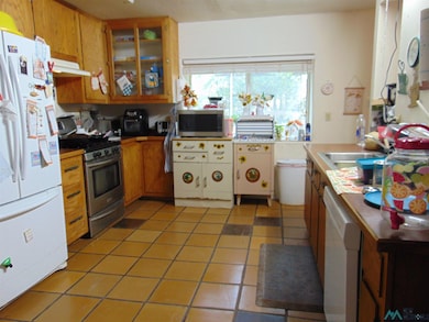 Kitchen with appliances with stainless steel finishes, brown cabinets, light tile patterned flooring, under cabinet range hood, and glass insert cabinets