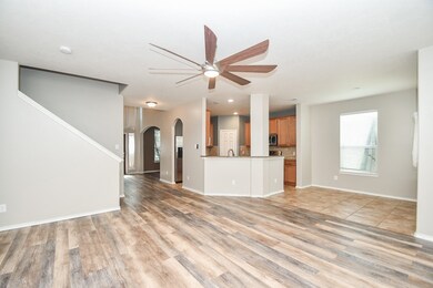 This is a spacious, open-plan living area featuring wood-look flooring, a modern ceiling fan, and plenty of natural light from multiple windows. The room flows seamlessly into a kitchen with wood cabinetry and a convenient breakfast bar.