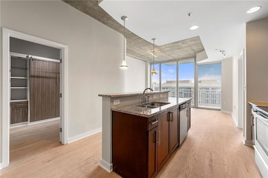 Kitchen with light wood-type flooring, dark brown cabinets, decorative light fixtures, a kitchen island with sink, and light stone counters