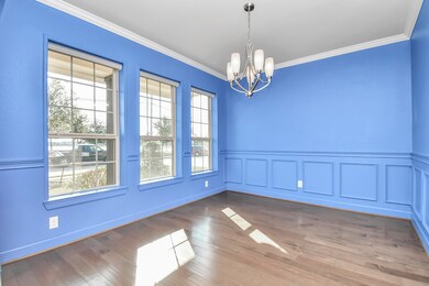 This is a bright formal dinning room featuring bold blue walls with white trim, wainscoting, three large windows for ample natural light, elegant hardwood floors, and a traditional chandelier.