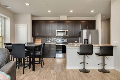 Kitchen with dark brown cabinetry, backsplash, light stone countertops, light wood-type flooring, and appliances with stainless steel finishes