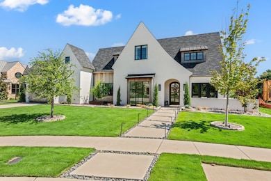 The exterior of the home blends modern elegance with timeless design, featuring smooth white stucco, steep gabled rooflines, and natural wood accents that add warmth and texture. A wide stone-lined walkway leads to a charming arched entry, flanked by tall, manicured greenery and custom iron lanterns. Black-framed windows and a mix of vertical siding and smooth finishes give the façade depth and character, setting the tone for the thoughtful design that continues throughout the interior