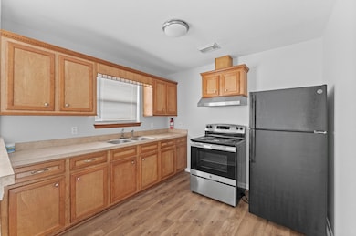 This kitchen features light wood cabinetry, a stainless steel oven with a range hood, and a black refrigerator. It has a double sink, ample countertop space, and wood-like flooring, creating a warm and functional cooking area.