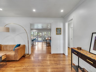 A bright open living space with hardwood floors, white walls, and a modern design. The area features a comfortable seating area and leads to a dining area with large windows offering a view outside.