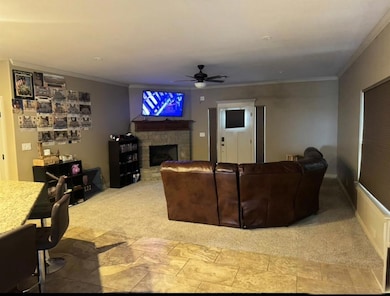 Carpeted living room featuring ornamental molding, a stone fireplace, and a ceiling fan