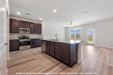 Kitchen with tasteful backsplash, dark brown cabinets, stainless steel appliances, light stone counters, and an island with sink