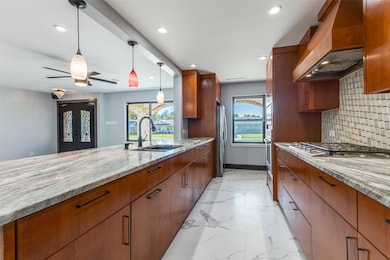 Kitchen featuring light stone countertops, brown cabinets, decorative backsplash, custom exhaust hood, and decorative light fixtures