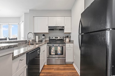 Kitchen with black appliances, under cabinet range hood, dark wood finished floors, white cabinets, and light stone countertops