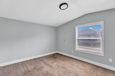 This is a cozy room with gray walls, a carpeted floor, and a large window providing natural light. The ceiling features a modern light fixture, and white trim complements the clean, simple design.