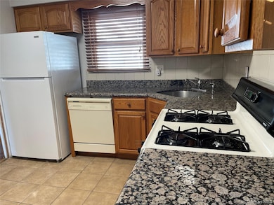 Kitchen featuring brown cabinets, white appliances, light tile patterned floors, decorative backsplash, and dark stone countertops