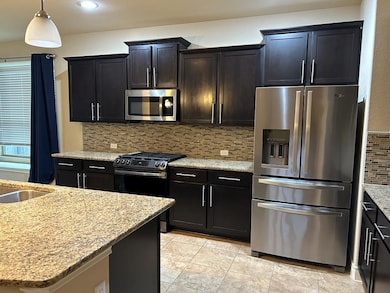 Kitchen with stainless steel appliances, backsplash, light stone countertops, dark cabinetry, and hanging light fixtures