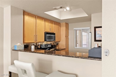 Kitchen with a textured wall, dark stone counters, appliances with stainless steel finishes, a textured ceiling, and light brown cabinetry