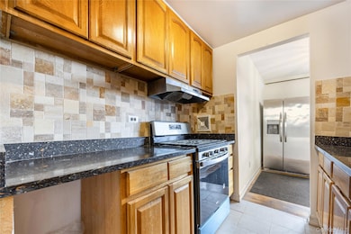Kitchen with stainless steel appliances, tasteful backsplash, brown cabinetry, light tile patterned floors, and under cabinet range hood